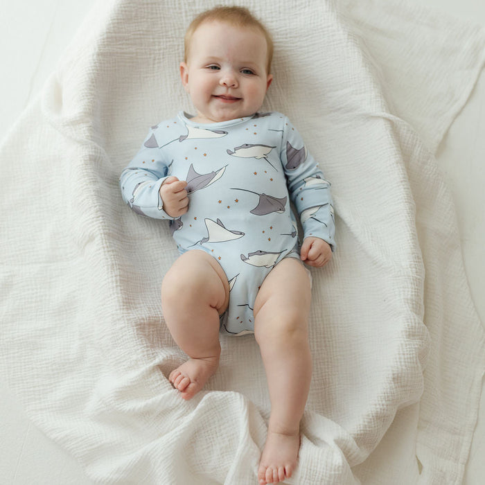 A baby lying on a white textured muslin blanket, wearing a light blue long-sleeve bodysuit with a gray and white whale print.
