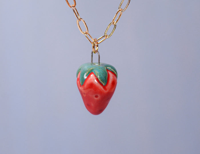 A ceramic pendant necklace with a strawberry design, featuring a red base with green leaves, attached to a gold-plated chain against a blue background.