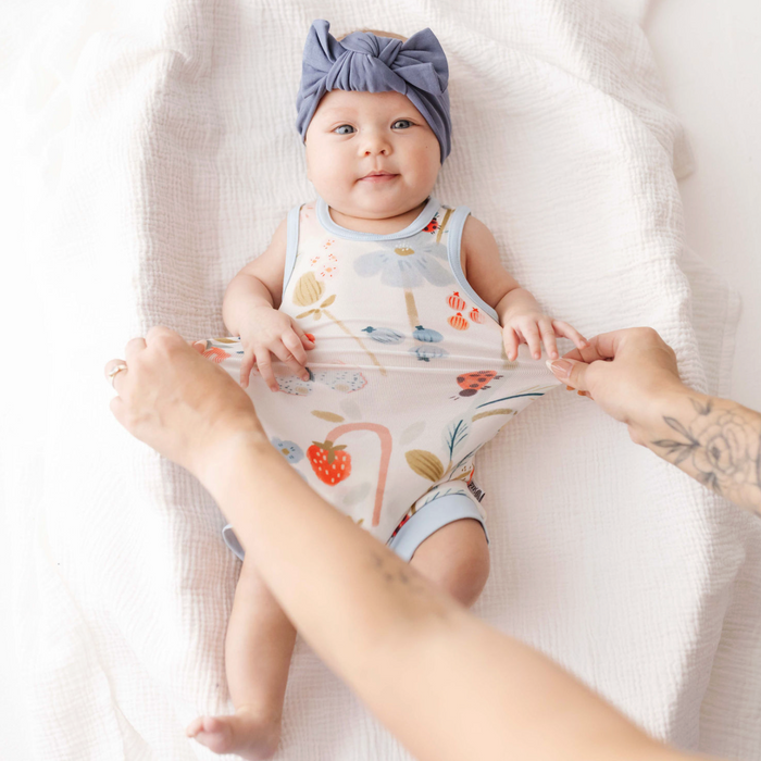 Baby wearing a floral romper and headband, lying on a white blanket with a person's hands holding the romper.