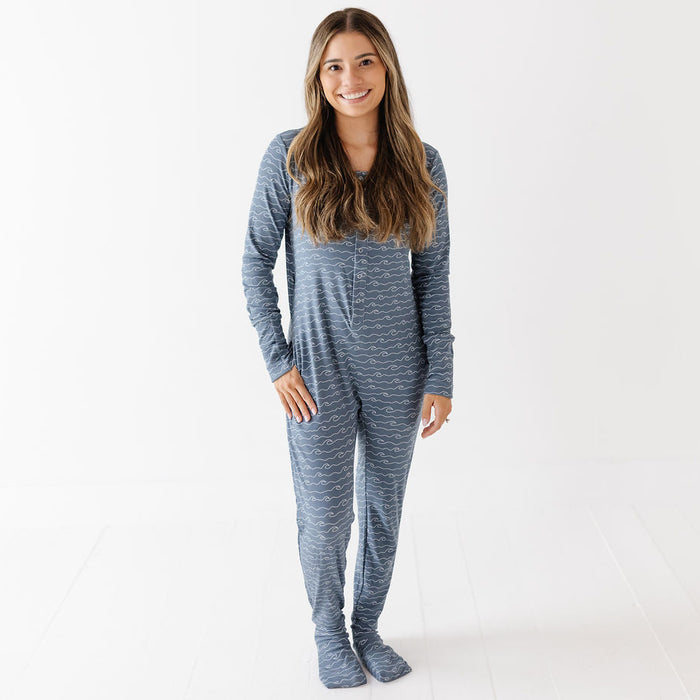A woman modeling a long-sleeve, one-piece footed pajama set with a heathered blue and gray pattern, standing against a clean white studio background.
