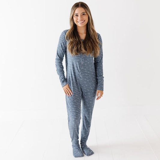 A woman modeling a long-sleeve, one-piece footed pajama set with a heathered blue and gray pattern, standing against a clean white studio background.