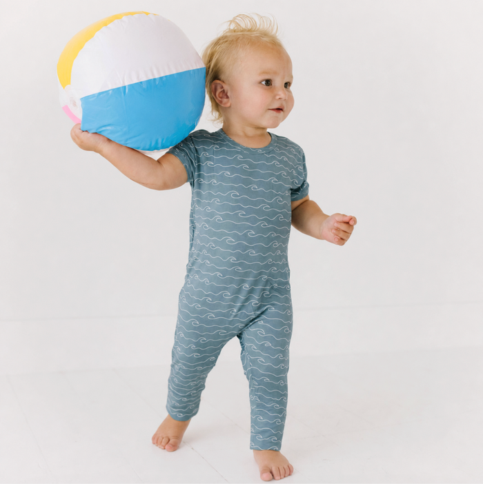 A toddler standing barefoot and holding a beach ball, wearing a short-sleeve one-piece jumpsuit in muted blue-gray with a white wavy line pattern. The background is a clean white studio setting.