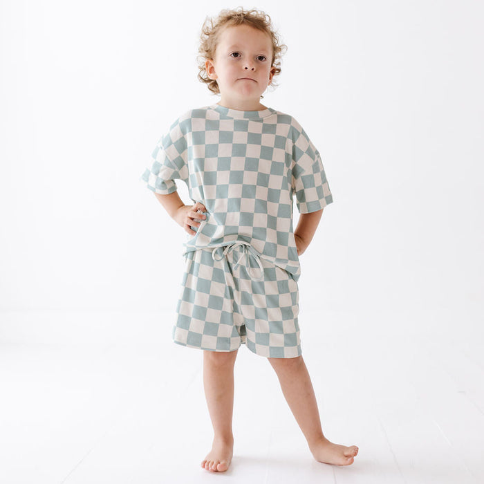 A toddler stands barefoot against a clean white studio background, wearing a matching short-sleeve top and shorts set in a pale blue and cream checkerboard pattern.