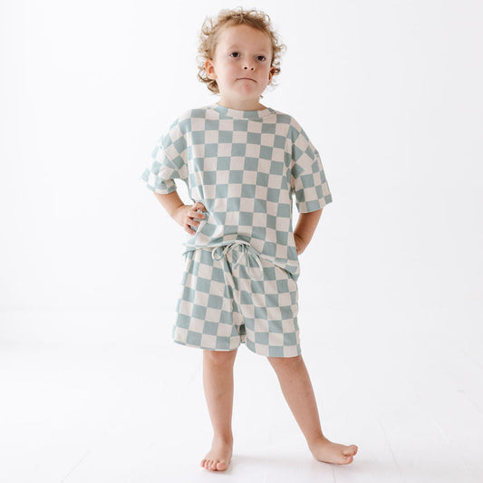 A toddler stands barefoot against a clean white studio background, wearing a matching short-sleeve top and shorts set in a pale blue and cream checkerboard pattern.