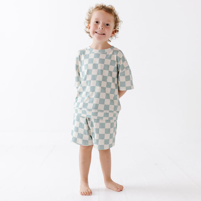 A young child with curly blonde hair stands barefoot against a clean white studio background, wearing a matching short-sleeve top and shorts set in a pale blue and off-white checkerboard pattern.