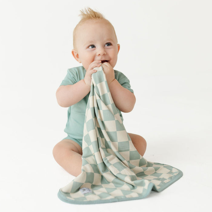 A baby sitting on a white studio floor, holding and chewing on a soft checkered cloth teether or pacifier clip in sage green and cream.