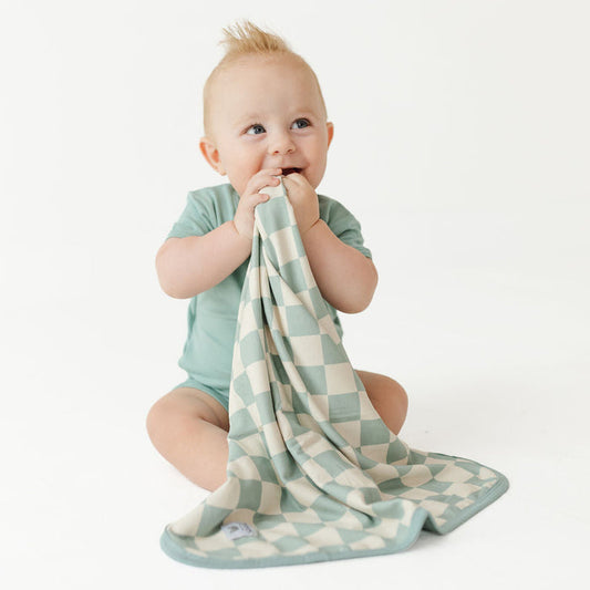 A baby sitting on a white studio floor, holding and chewing on a soft checkered cloth teether or pacifier clip in sage green and cream.