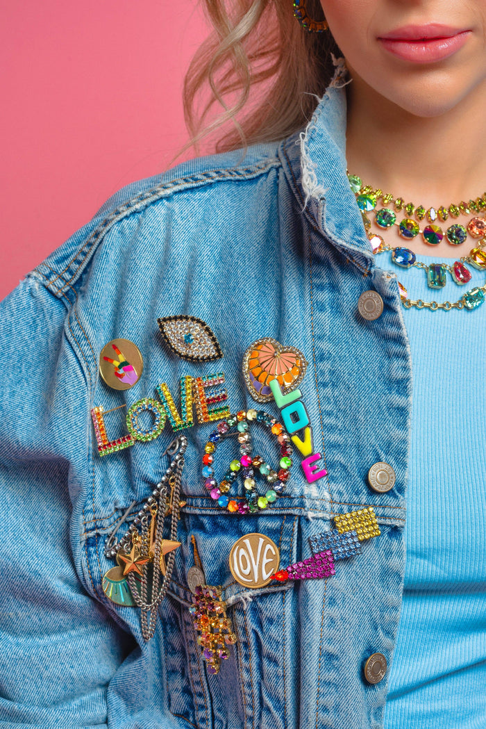Person wearing a denim jacket with colorful patches and accessories against a pink background