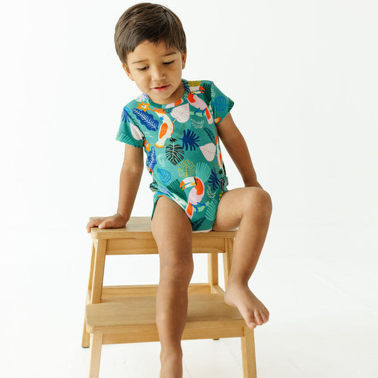 A young boy wearing a short-sleeve tropical-print baby bodysuit sits on a small wooden step stool against a clean white background.