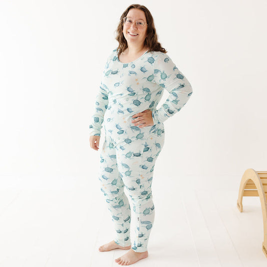 A woman wearing a long-sleeve, one-piece pajama set with a blue and green leaf pattern on a white background, standing in a bright studio setting.