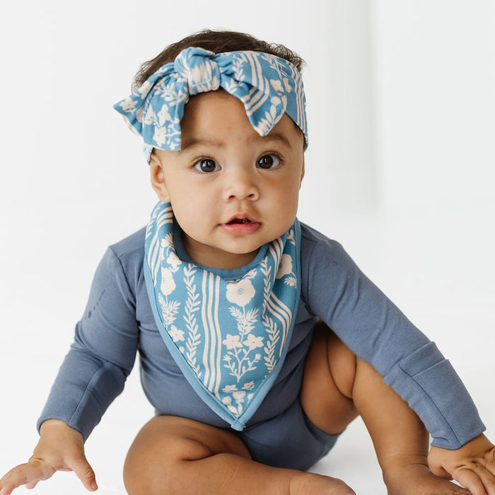 A baby wearing a slate blue long-sleeve bodysuit, a matching blue and white patterned bib, and a matching blue and white patterned headband, sitting on a white studio floor.