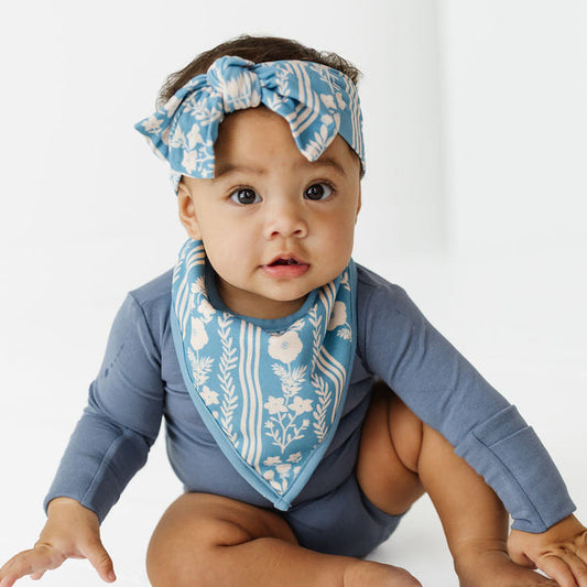 A baby wearing a slate blue long-sleeve bodysuit, a matching blue and white patterned bib, and a matching blue and white patterned headband, sitting on a white studio floor.