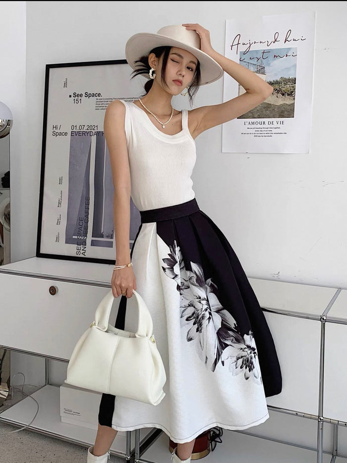 Woman in a white sleeveless top and black floral skirt holding a white handbag, standing in a room with posters on the wall.