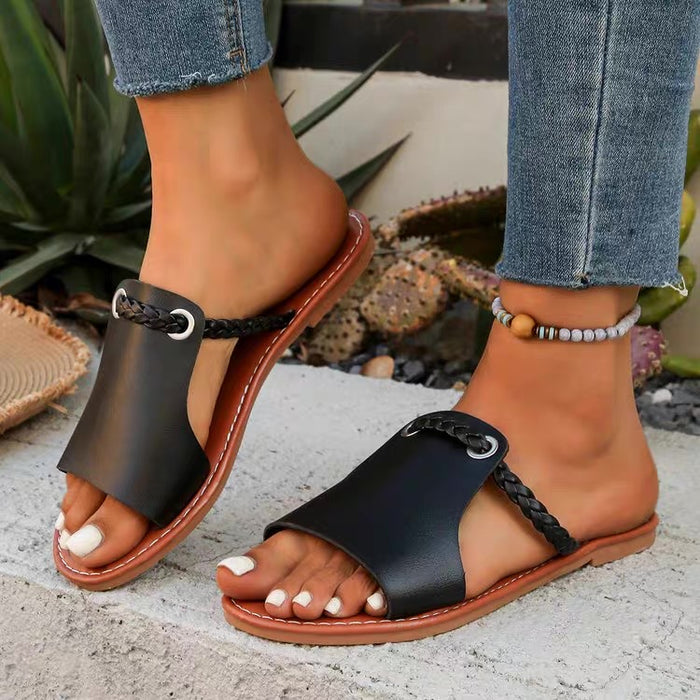 Black sandals with brown soles worn by a person standing on a stone surface with plants in the background.