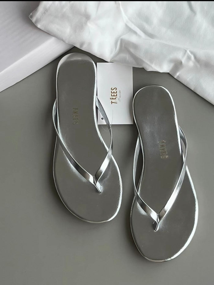 Silver flip-flops on a gray surface with a white cloth and 'TEES' box in the background.