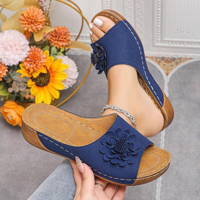 Blue sandals with floral design held by a hand, with flowers and a decorative wall in the background.