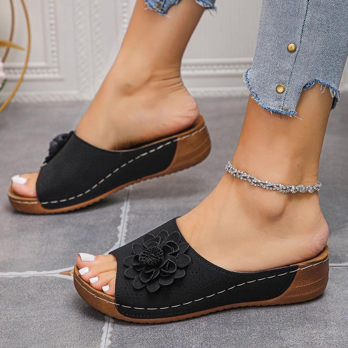 Black sandals with flower detail worn by a person on a gray floor.