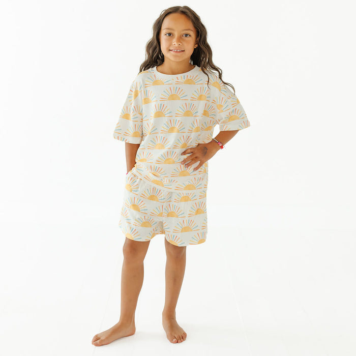 A young girl with long brown hair stands barefoot against a plain white studio background, wearing a short-sleeve white nightgown or pajama dress with a repeating yellow sunburst pattern.