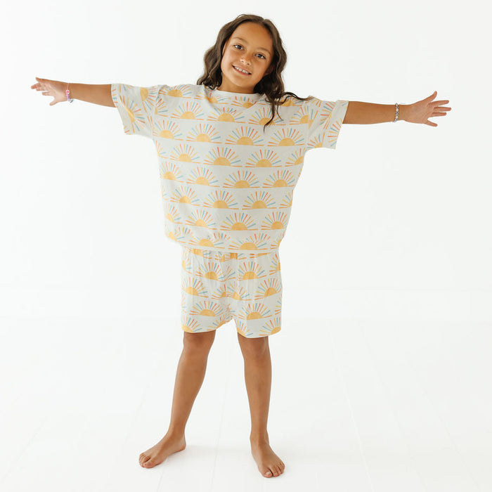 A young girl stands barefoot with her arms outstretched, wearing a matching two-piece pajama set featuring a white base with a yellow sunburst pattern. The set includes a short-sleeve top and matching shorts, photographed against a seamless white studio background.