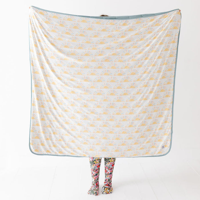 A person holds up a large square blanket or throw with a white background, yellow sunburst pattern, and a muted blue border. The person is only visible at the bottom through floral socks, against a clean white studio background.
