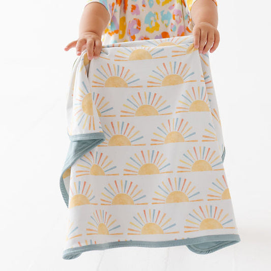 A toddler wearing a colorful patterned shirt and a purple headband holds up a reusable cloth baby blanket with a yellow sunburst pattern and a blue border against a white studio background.